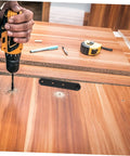 Concealed hinge plate (stainless steel) with three mounting holes on wooden workbench; drill and tools nearby.