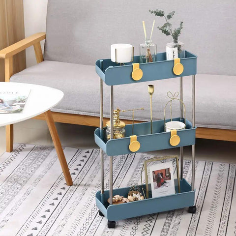 Navy blue three tier slim mobile shelving unit on wheels with slide out storage cart featuring decor items in a living room corner.