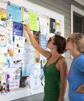 Two women using Top Shelf Bulk Push Pins For Bulletin Boards And Cork Walls on a busy bulletin board.