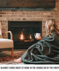 Person wrapped in olive green Beefy Warmth And Rugged Durability Military Wool Blanket for outdoors, sitting by a fireplace.