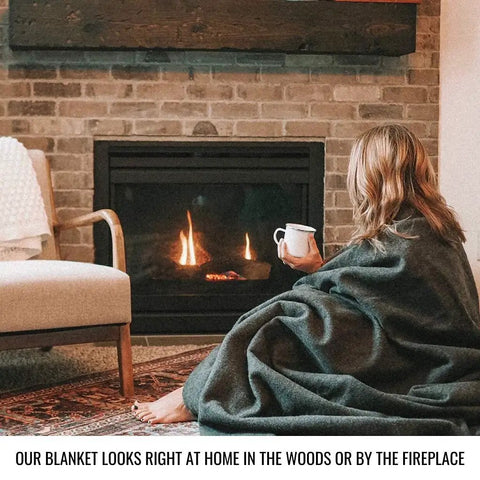Person wrapped in olive green Beefy Warmth And Rugged Durability Military Wool Blanket for outdoors, sitting by a fireplace.