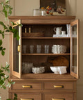 Open wooden kitchen cabinet with glassware, plates, and cutting board; antique brass cup pulls visible on drawers below.