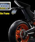 Carbon Fiber Motorcycle License Plate Frame With Stainless Steel Tag Holder Bracket And Included Screws Nuts Caps 1 Pack For Moto Moped Motorbike Scooter Bike mounted on a motorcycle with visible rear wheel and suspension.