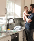 Man holding baby in kitchen with Delta Touch2O faucet, featuring VoiceIQ Activation Module for Touch2O Kitchen Faucets Compatible with Alexa and Google Assistant Voice Control Smart Home Upgrade Kit EP100855 Easy Setup and Hands Free Kitchen Control.