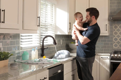 Man holding baby in kitchen with Delta Touch2O faucet, featuring VoiceIQ Activation Module for Touch2O Kitchen Faucets Compatible with Alexa and Google Assistant Voice Control Smart Home Upgrade Kit EP100855 Easy Setup and Hands Free Kitchen Control.