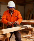 Worker in hard hat and safety goggles cutting wood with chainsaw in workshop