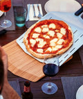 14 Inch Perforated Pizza Peel With Long Handle Aluminum Lightweight Pizza Paddle holding a pizza on a dining table.