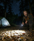 Man using Compact LED Mini Flashlight With Tiny But Mighty Brightness in forest campsite at night, lighting path, tent nearby