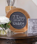Decorative setup with a round chalkboard plate displaying '7 Days Until Our Wedding' next to a vase of white roses and a small sign.