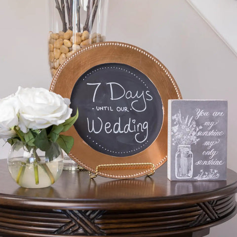 Decorative setup with a round chalkboard plate displaying '7 Days Until Our Wedding' next to a vase of white roses and a small sign.