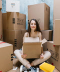 Person sitting amongst boxes with an open cardboard box, beside a stack of Waterproof Bubble Mailers 25 Pack 28x37 cm Plus 6 cm Thick Self Adhesive Padded Envelopes for Tough As Nails Shockproof Shipping and Mailing, Lightweight Packaging Bags for Fragile Items.