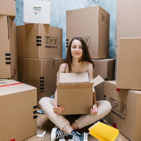 Person sitting amongst boxes with an open cardboard box, beside a stack of Waterproof Bubble Mailers 25 Pack 28x37 cm Plus 6 cm Thick Self Adhesive Padded Envelopes for Tough As Nails Shockproof Shipping and Mailing, Lightweight Packaging Bags for Fragile Items.