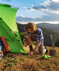 Person setting up green tent with Lightweight 18.5 Inch Folding Shovel For Camping And Hiking beside them, mountains in background.