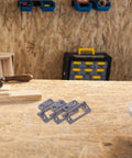Front view: ten stainless steel cabinet hinge plates with screw holes on a wooden workbench