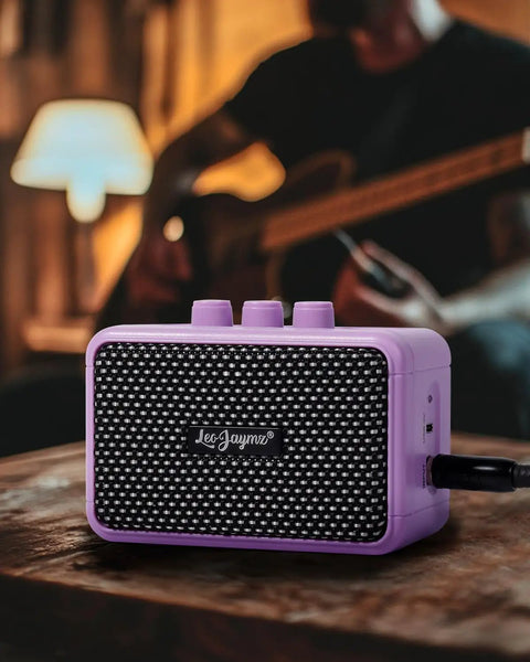Compact 5W Guitar Amp For On The Go With Dual Speakers Bluetooth Wireless Connectivity Rechargeable Battery Clean And Overdrive Modes Gain Tone And Volume Controls Headphone Jack Ideal For Practice And Small Gigs Purple on table with blurred guitarist in background.