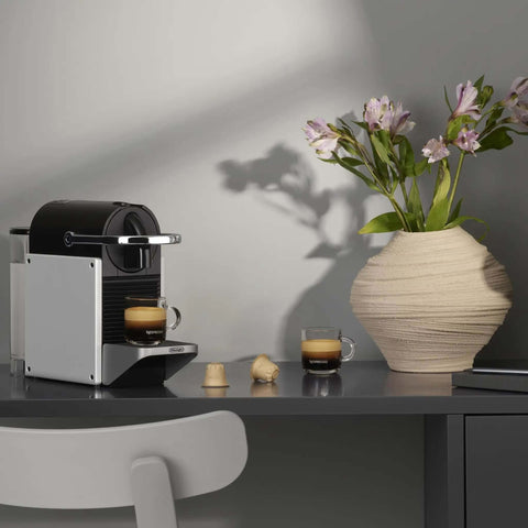 Pixie Aluminum Espresso Machine For Capsule Coffee Lovers on a countertop, brewing espresso, beside a vase of flowers.