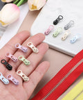 Various colored zipper sliders for 30 Pack Zipper Sliders Size Three Metal Repair Replacement With Zip Pull No Sewing Needed For Bags Wallets Jackets And Closed End Zippers Purple Great For DIY Repairs And Everyday Fixes displayed on a surface with a hand holding some