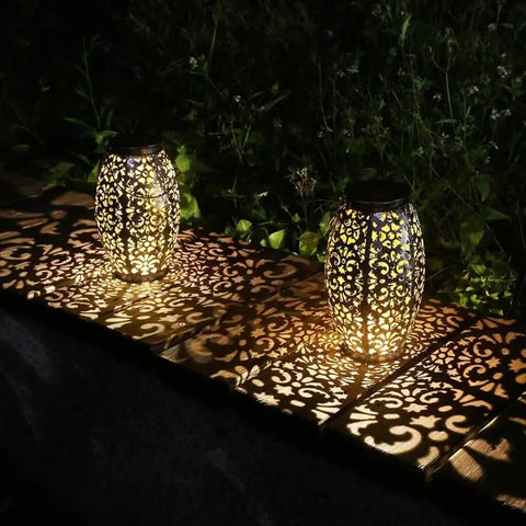 Two cool solar lantern lights for hanging or table outdoor night glow for patio courtyard garden with unique patterned hollowed design and warm ambient light, weatherproof, creating shadows on a wooden surface.
