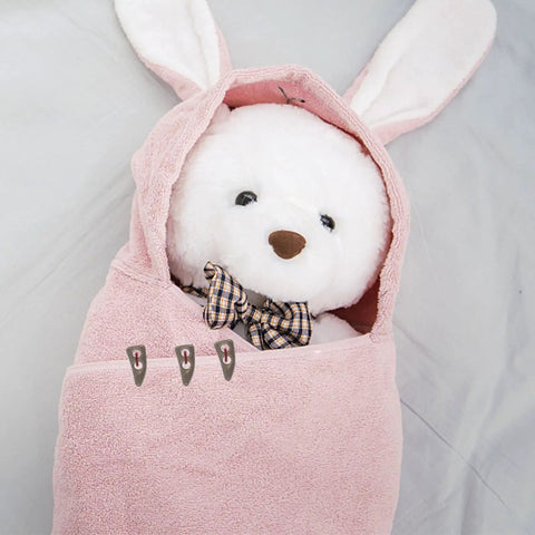 Stuffed animal wrapped in pink bunny-ear blanket with a plaid bow tie, fastened with clips, on a gray background.