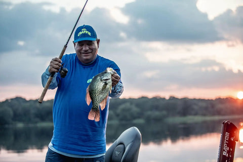 Person holding a crappie fish with a fishing rod at sunset on a lake.
