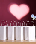 White Kraft Paper Gift Bags With Handles Pack Of 25 Medium 8 By 4.5 By 10 Inches on display against a heart backdrop.