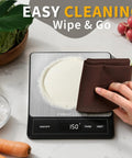 Rechargeable Digital Kitchen Scale Your Go To Helper For Home Cooks With 33 Lb Capacity And 1g And 0.05oz Precision Type C Stainless Steel LCD Display Switchable Grams Ounces For Cooking Baking Meal Prep Ash Silver being wiped clean with vegetables in background.