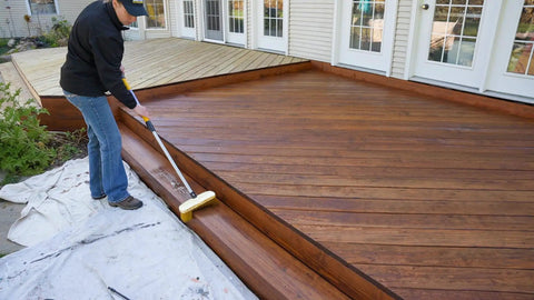 Person applying Heavy Duty Wood Stain And Sealer In One For Decks, 5 Gallon Semi Transparent Waterproof Coating In Light Walnut.