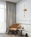 Three Light Modern Wall Sconce With Clear Glass Globe And Gold Base For Bedside Or Living Room Sleek Gold Wall Lamp Delivers Warm Ambient Glow And Contemporary Decor, mounted by a tufted armchair.