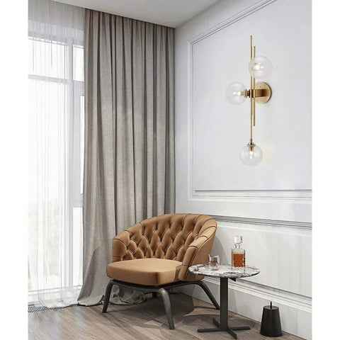 Three Light Modern Wall Sconce With Clear Glass Globe And Gold Base For Bedside Or Living Room Sleek Gold Wall Lamp Delivers Warm Ambient Glow And Contemporary Decor, mounted by a tufted armchair.