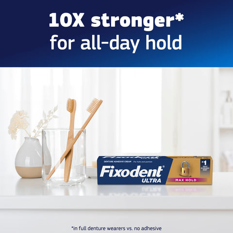 Box of Ultra Max Hold Denture Adhesive next to toothbrushes on bathroom counter with text '10X stronger for all-day hold'