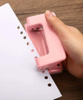 Hand holding pink paper puncher creating clean holes in sheet, showcasing durable design and ease of use for home and office.