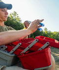 Person using a Three-Tray Tackle Box With Cantilever Design For Easy Access Large Portable Fishing Lure Storage Organizer With 32 Compartments And Removable Dividers Flip-Top Lid For Accessories Red And Gray by a lake.