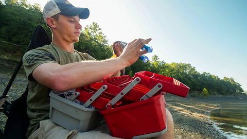 Person using a Three-Tray Tackle Box With Cantilever Design For Easy Access Large Portable Fishing Lure Storage Organizer With 32 Compartments And Removable Dividers Flip-Top Lid For Accessories Red And Gray by a lake.