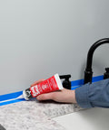 Applying Kwik Seal Kitchen And Bath Adhesive Caulk White 2.8 Oz Waterproof Sealant along a bathroom sink edge with a blue tape guide.