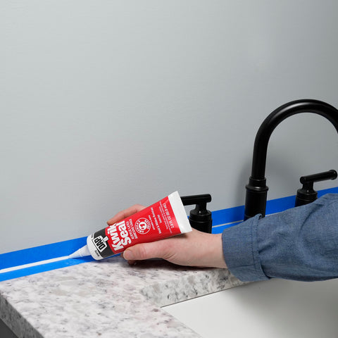 Applying Kwik Seal Kitchen And Bath Adhesive Caulk White 2.8 Oz Waterproof Sealant along a bathroom sink edge with a blue tape guide.