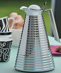 Glass Vacuum Copper Carafe With Chrome Plated Spout And Stainless Steel Handle Keeps Hot For 12 Hours And Cold For 24, 1.0 L Capacity, Dope Chrome Finish, Insulated Beverage Server For Hot And Cold Drinks on a table with cups in the background.