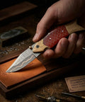 Hand holding Handmade Copper Damascus Folding Knife With Ladder Pattern Blade Brass Engraved Red Resin Handle Leather Sheath