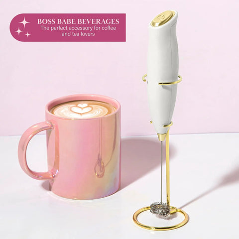 Electric Handheld Milk Frother With Double Coil Whisk And Gold Stand next to a pink mug with latte art.