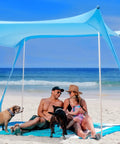 Beach Tent Sun Shelter UPF50 Plus Easy Pop Up Canopy providing shade for a couple and dogs on a sandy beach.