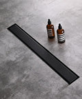 Black Sleek 36 Inch Linear Shower Floor Drain Kit in 304 Stainless Steel installed on grey tile floor with bottles nearby.