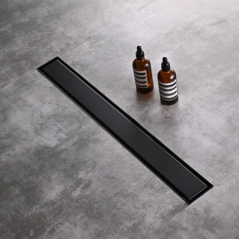 Black Sleek 36 Inch Linear Shower Floor Drain Kit in 304 Stainless Steel installed on grey tile floor with bottles nearby.