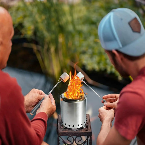 Two people roasting marshmallows over a Pellet Tabletop Fire Pit 7 Inch Mini Outdoor Fire Pit With Removable Ash Pan Carry Bag Marshmallow Roasting Sticks Loading Spoon Ideal For Patio Camping Backyard Picnics on a patio.