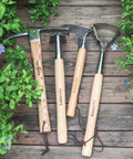 All In One Double Sided Garden Rake And Cultivator Tool With Wood Handle on wooden deck surrounded by greenery