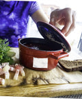 Person opening a red casserole dish on a wooden board with herbs, meat slices, and bread