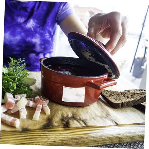 Person opening a red casserole dish on a wooden board with herbs, meat slices, and bread