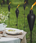 Black Six Pack Citronella Tiki Torches Outdoor 59.5 Inch Metal torches with 3-prong stakes in garden near dining table.