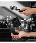 Perfect Your Daily Grind With This Top Shelf Height Adjustable Coffee Tamping Station Built For 58mm Portafilters And Equipped With A Thick Silicone Base, featuring a hand holding a portafilter above a black tamping station.