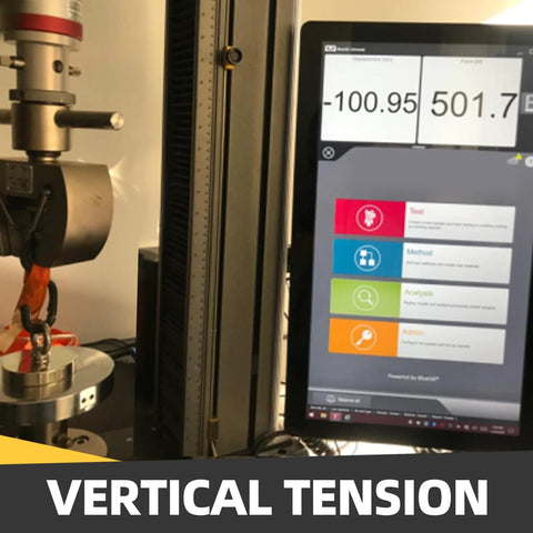 Vertical tension testing rig with metal clamp and hanging ring; computer monitor displays measurement readouts.