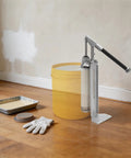 Heavy Duty Manual Loading Paste Pump With 15 Inch Handle And Stable Aluminum L Base For Thick Liquids beside a yellow bucket in home setting.