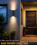 Modern Outdoor Wall Lights Up and Down Cylinder Sconce Matte Black illuminating entrance with plants and tiled pathway.